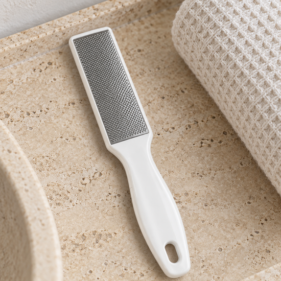 Dual-sided stainless steel pedicure foot rasp and callus remover file with ergonomic white handle on stone vanity tray