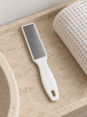 Dual-sided stainless steel pedicure foot rasp and callus remover file with ergonomic white handle on stone vanity tray