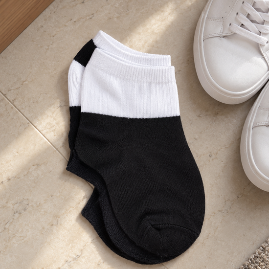 Pair of men's low cut ankle athletic socks in two-tone contrast styling with white ribbed elastic cuff and solid black foot resting next to white low-top sneakers