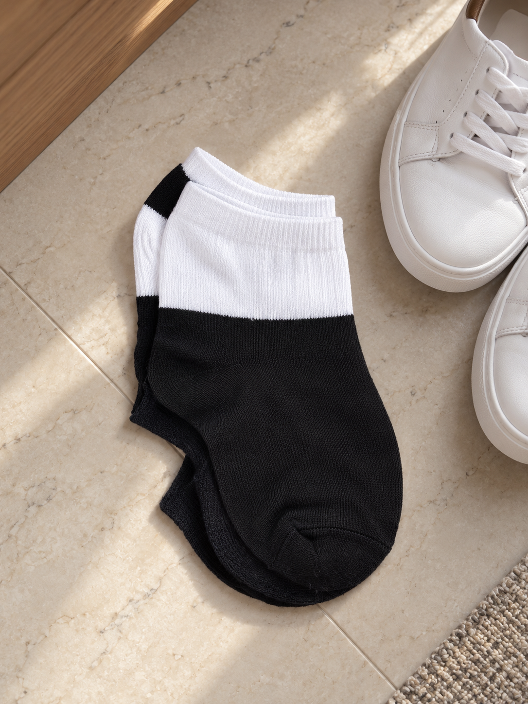 Pair of men's low cut ankle athletic socks in two-tone contrast styling with white ribbed elastic cuff and solid black foot resting next to white low-top sneakers
