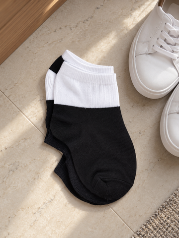 Pair of men's low cut ankle athletic socks in two-tone contrast styling with white ribbed elastic cuff and solid black foot resting next to white low-top sneakers