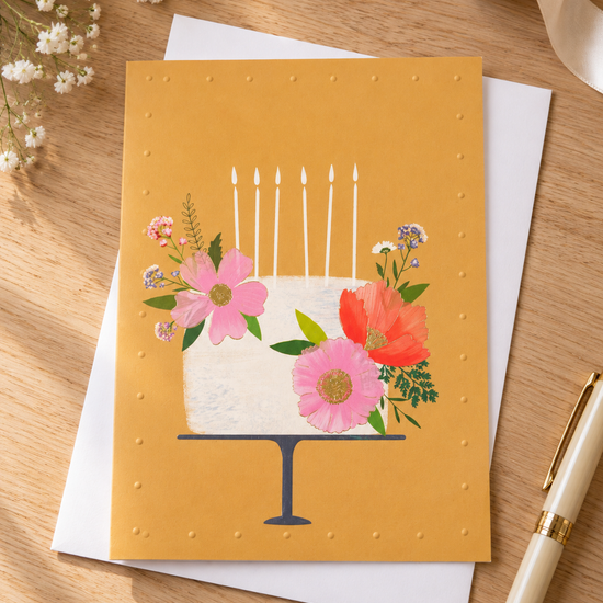 Artistic botanical floral birthday cake on navy stand with lit candles on warm yellow greeting card