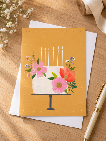 Artistic botanical floral birthday cake on navy stand with lit candles on warm yellow greeting card