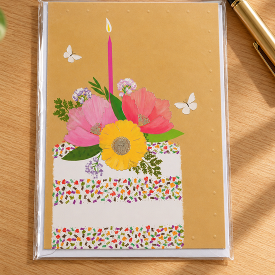 Colorful confetti sprinkle birthday cake with pink candle and floral topper on warm yellow greeting card